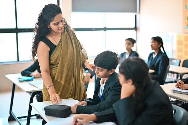 Female school teacher bends over a student’s desk to give individual guidance while children in uniforms work on their notebooks, showing a supportive learning environment in a bright classroom.