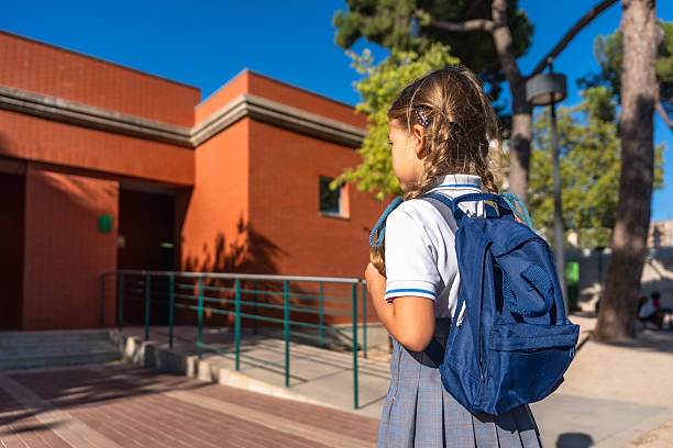 Young girl with braids and blue backpack walks toward school building on a sunny day, ready for class and the new school year, rear view, elementary student returning to campus