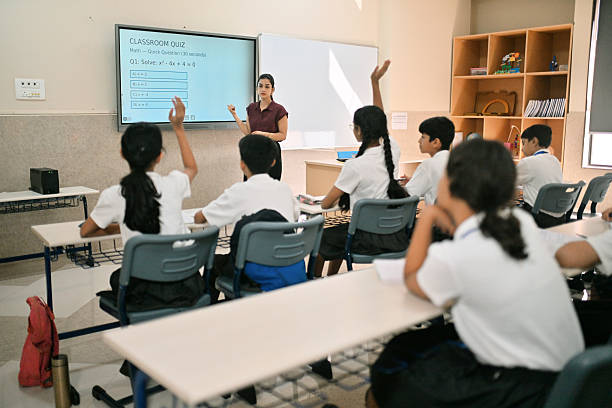 Group of Indian school students in uniform attending a math class, raising their hands to answer a question displayed on the smart screen. Female teacher stands near the board conducting a quick classroom quiz on quadratic equations. Concept of active learning, STEM education, student participation, and modern classroom environment in India.